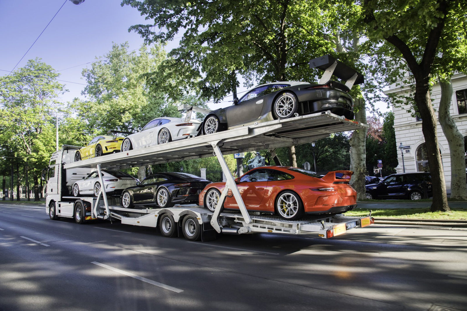 A car transporter truck is carrying six luxury sports cars, including the exclusive Porsche Collection, three on the upper deck and three on the lower, as it cruises down a tree-lined urban street.