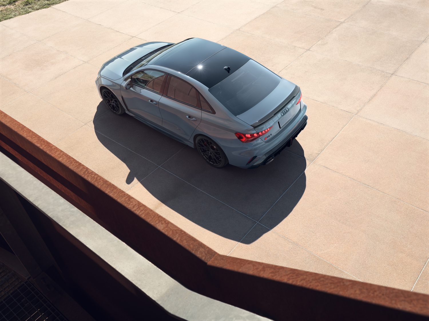 Blue sedan parked on a concrete surface, viewed from above. The car has a sunroof and black wheels, and it's casting a shadow on the ground. A metal railing is visible in the foreground.