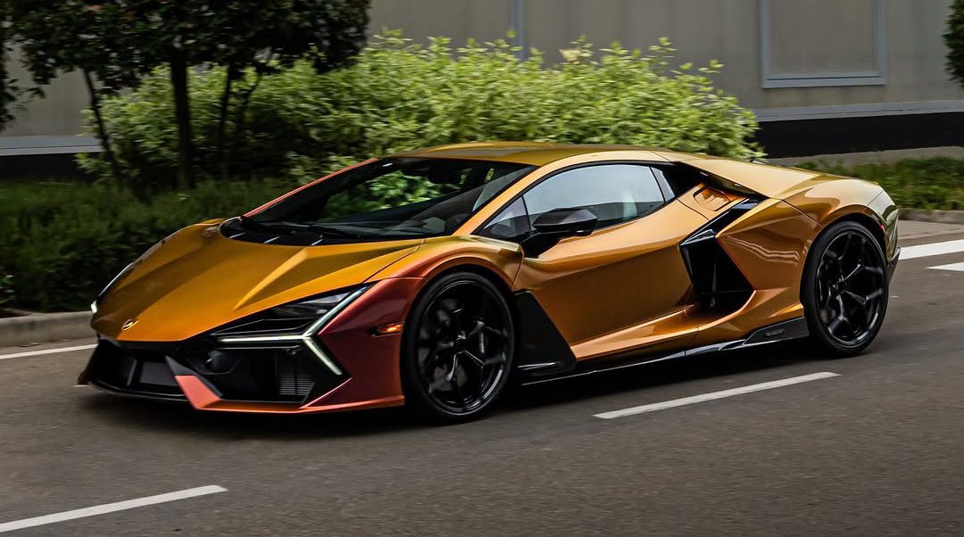 A sleek, Oro Alba-colored sports car with black accents drives on a road, surrounded by greenery. This stunning Revuelto model turns heads as it reflects the style and sophistication associated with Lamborghini Dallas.
