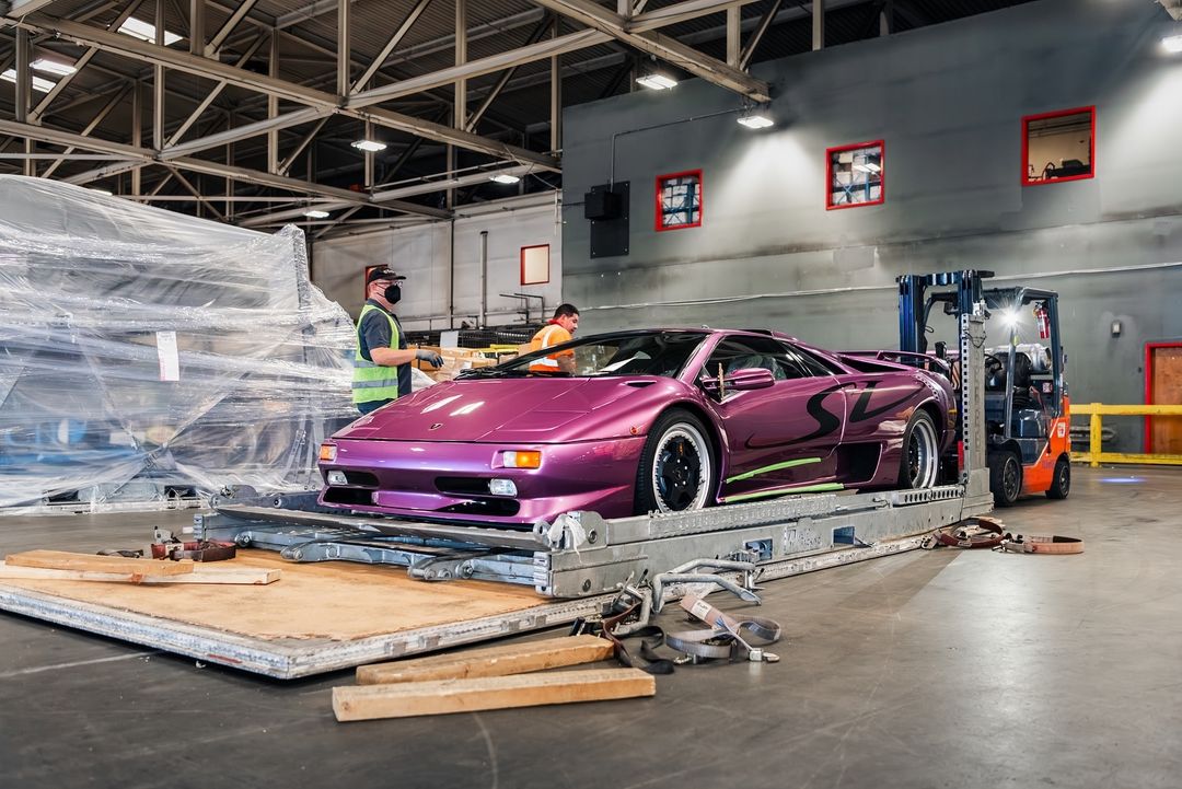 The Lamborghini Diablo SV in striking Viola 30th is positioned on a platform inside a CURATED warehouse. Nearby, two workers and a forklift stand ready to assist.