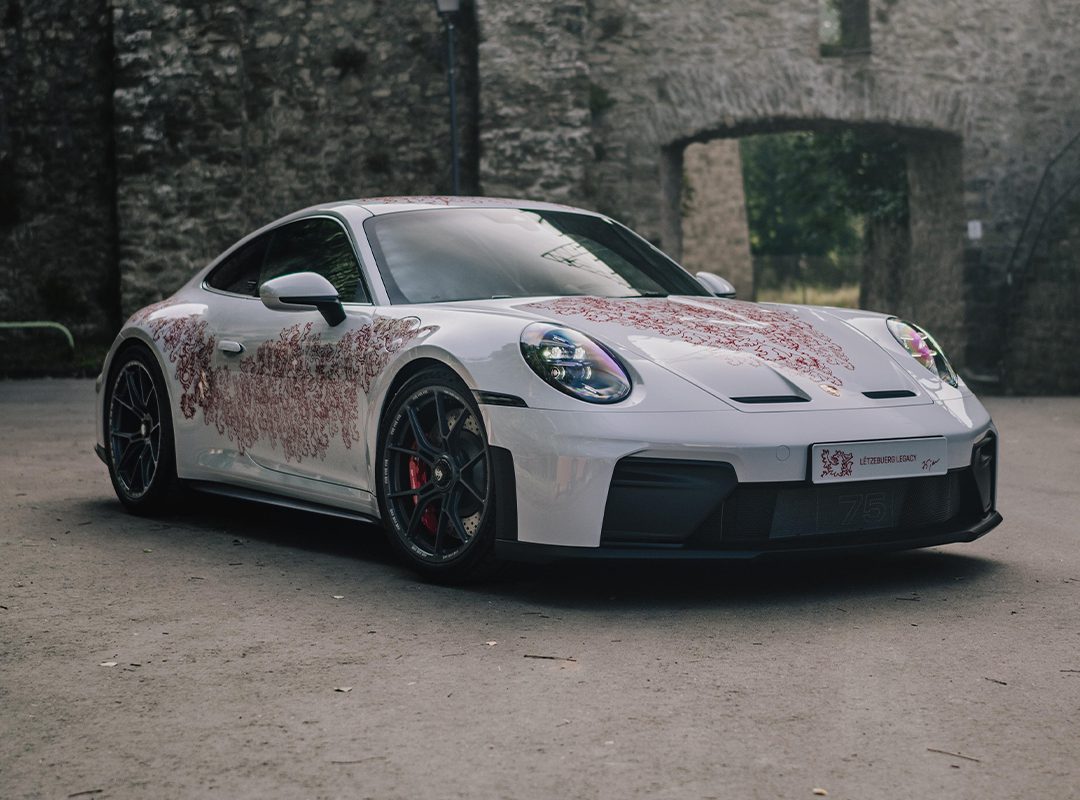 A Luxembourg-inspired Porsche Sonderwunsch 911 GT3 Touring with red floral patterns is parked on a paved surface in front of a stone wall with arched openings.