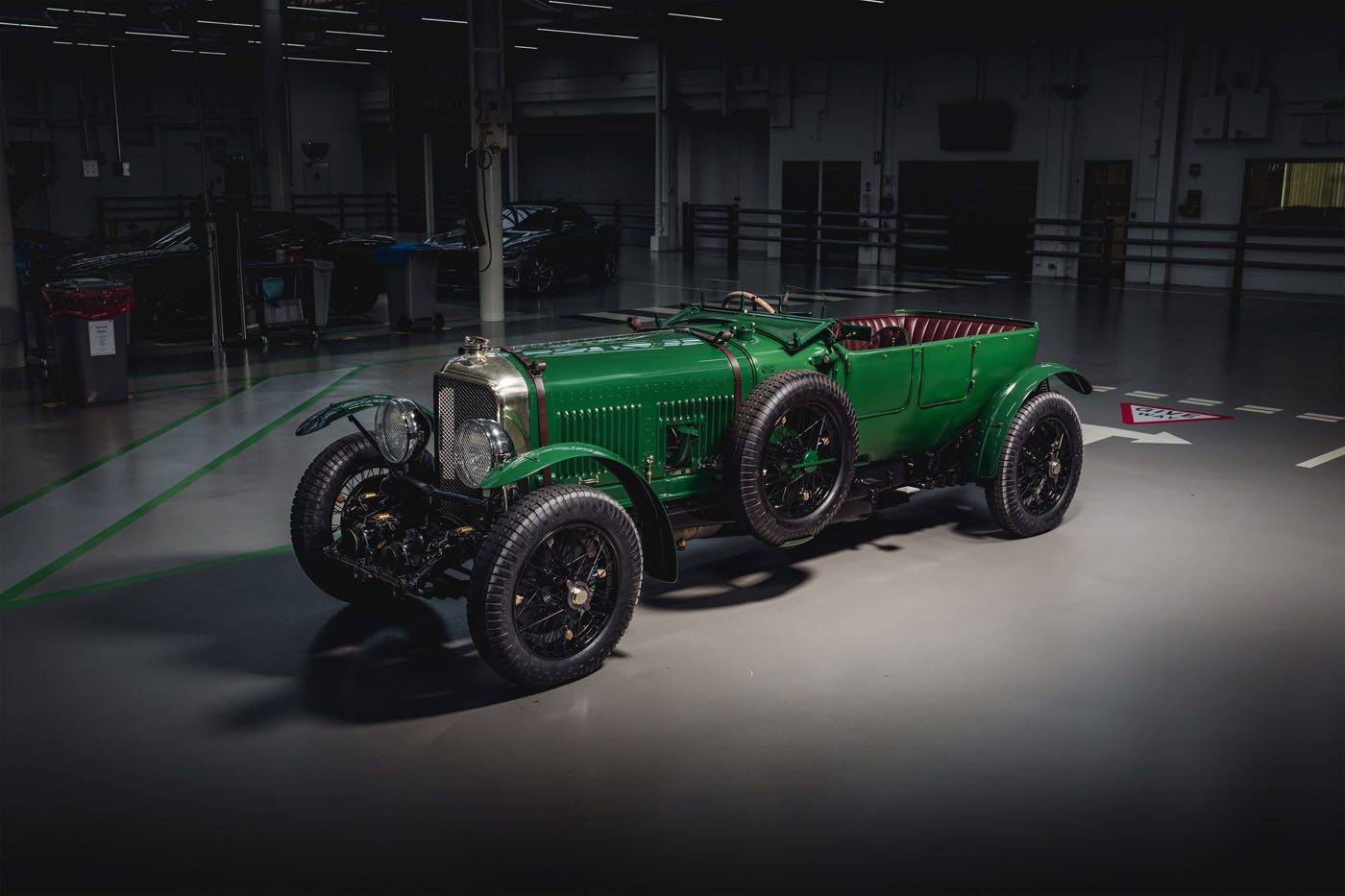 A vintage green Bentley, part of the Speed Six Continuation Series, stands parked indoors under dim lighting, showcasing classic design features and large spoked wheels with a luxurious red leather interior.