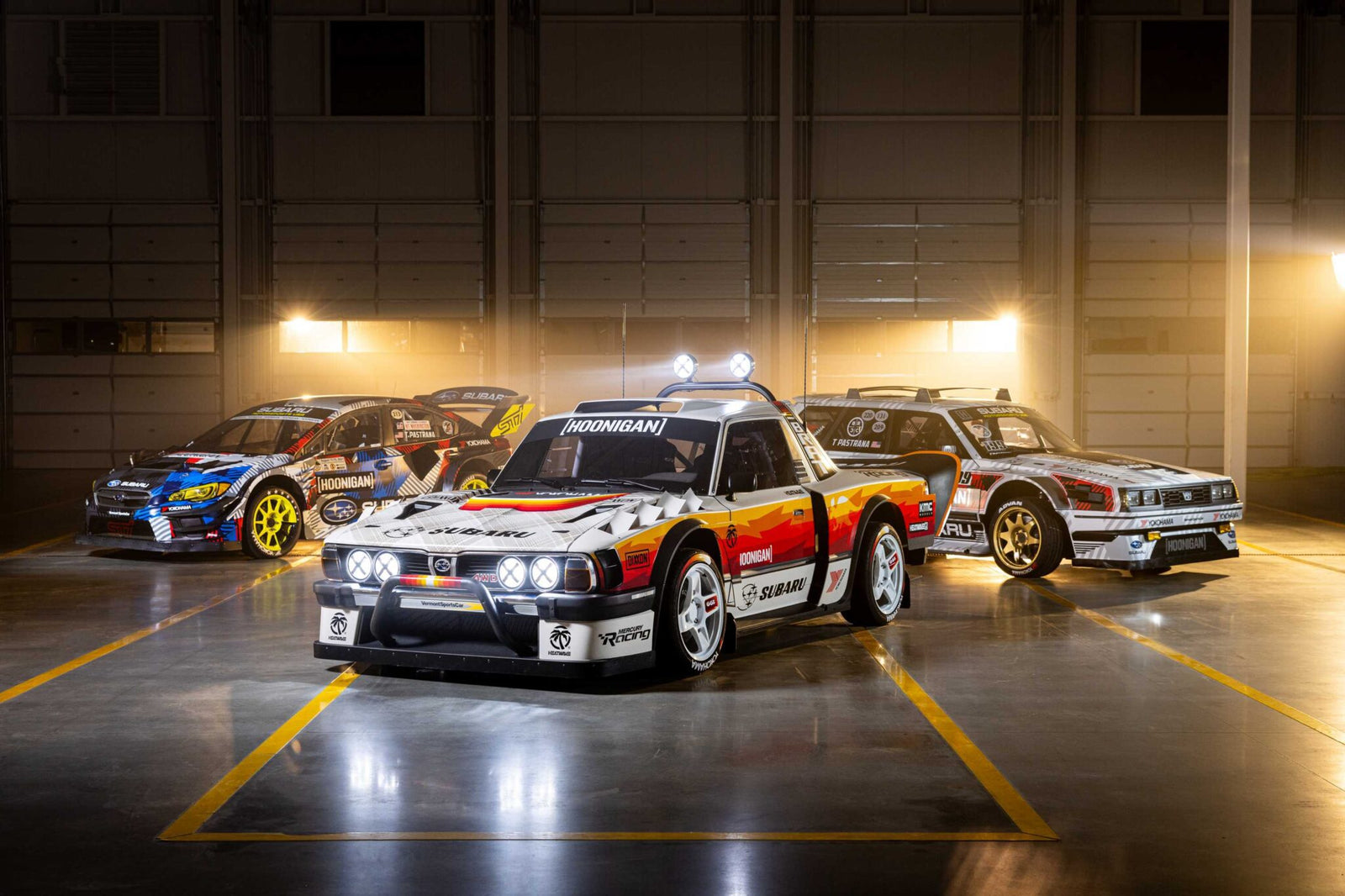 Three modified race cars, including the Subaru Brataroo, with decals and sponsor logos are parked indoors in a well-lit garage with a dark background, showcasing their Gymkhana-ready style for SEMA 2025.