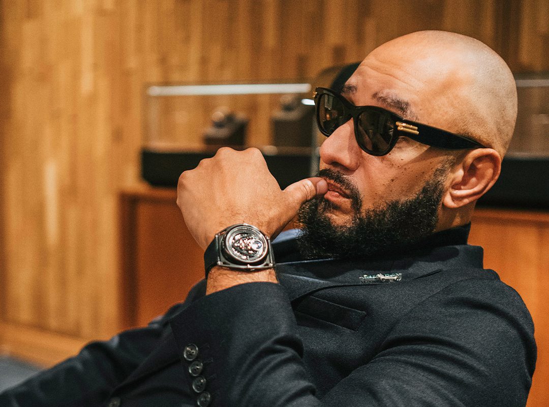 Bald man with sunglasses sits in a chair, hand on chin, exuding Swizz Beatz's style. He's wearing a black suit and a striking De Bethune GMT watch. Behind him, a wooden wall and blurred objects complete the scene.