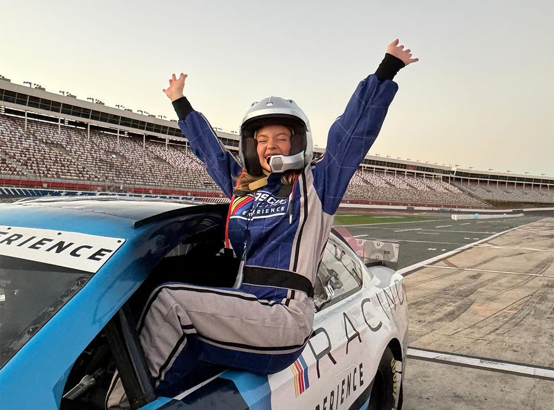 Person in a racing suit and helmet sits triumphantly on a NASCAR race car with arms raised in celebration at a racetrack, channeling the thrill of an unforgettable racing experience.