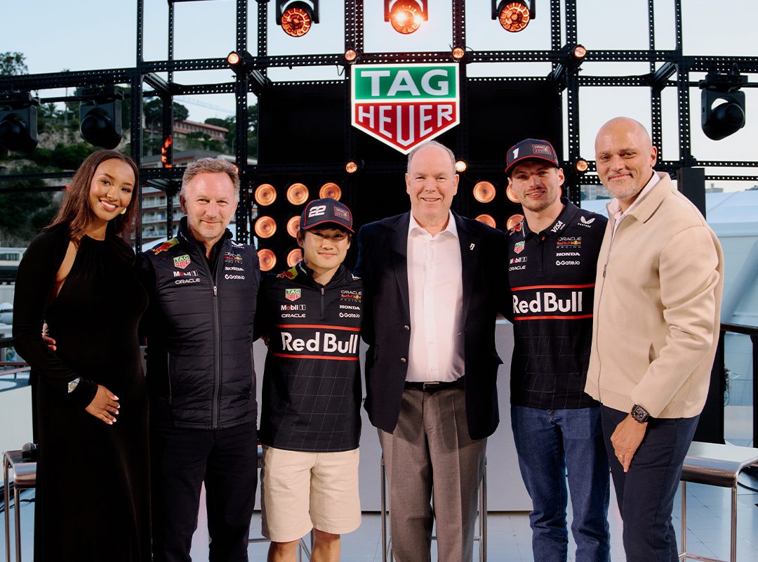 Six people pose together in front of a TAG Heuer sign, with two wearing Red Bull racing gear, at an outdoor Monaco Grand Prix event surrounded by lights and equipment.