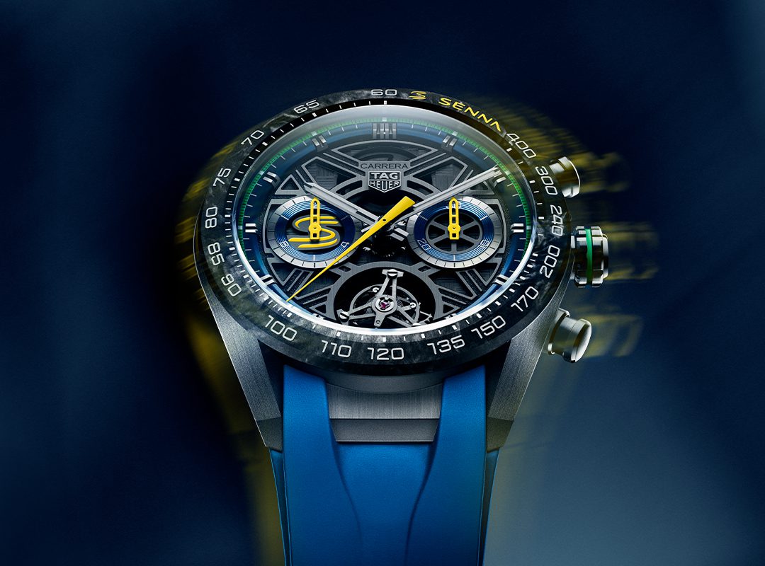 Close-up of a TAG Heuer Carrera Chronograph watch with a complex dial, visible mechanics, and a blue rubber strap on a blurred blue background.