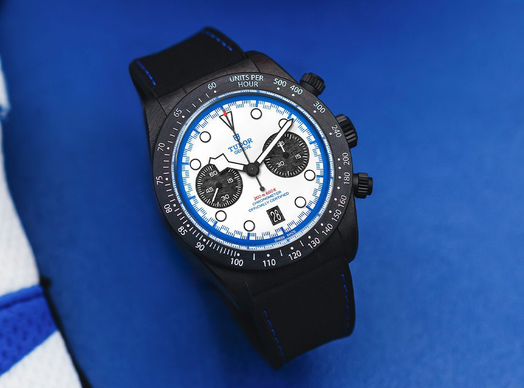A TUDOR Black Bay Chrono watch with a white and blue dial, black subdials, black bezel, and black strap is displayed on a blue surface—an eye-catching tribute to the Miami F1 spirit.