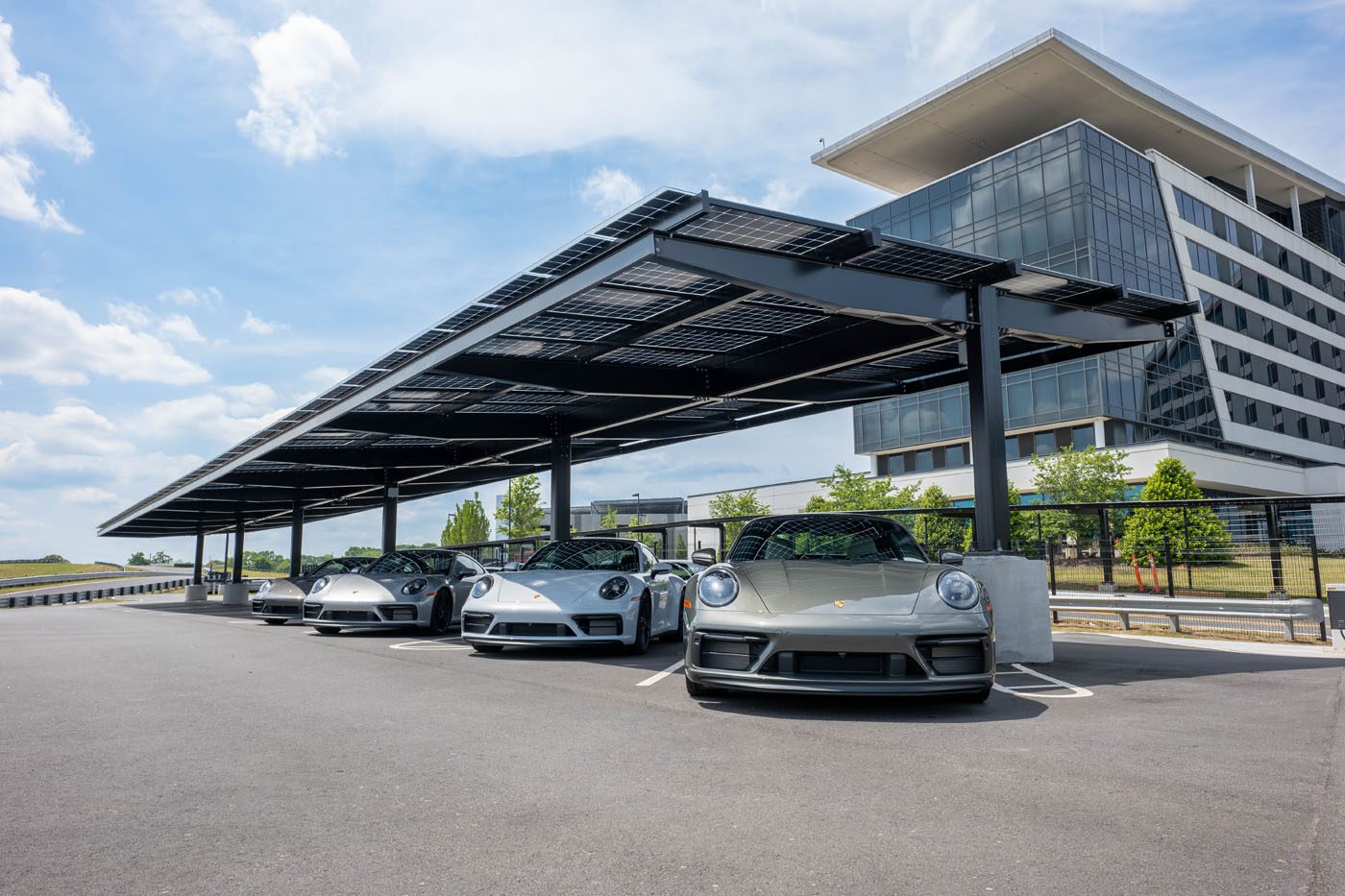 Four gray Porsche sports cars are parked under a large solar panel canopy at One Porsche Drive in Atlanta, beside a modern glass office building on a clear day.