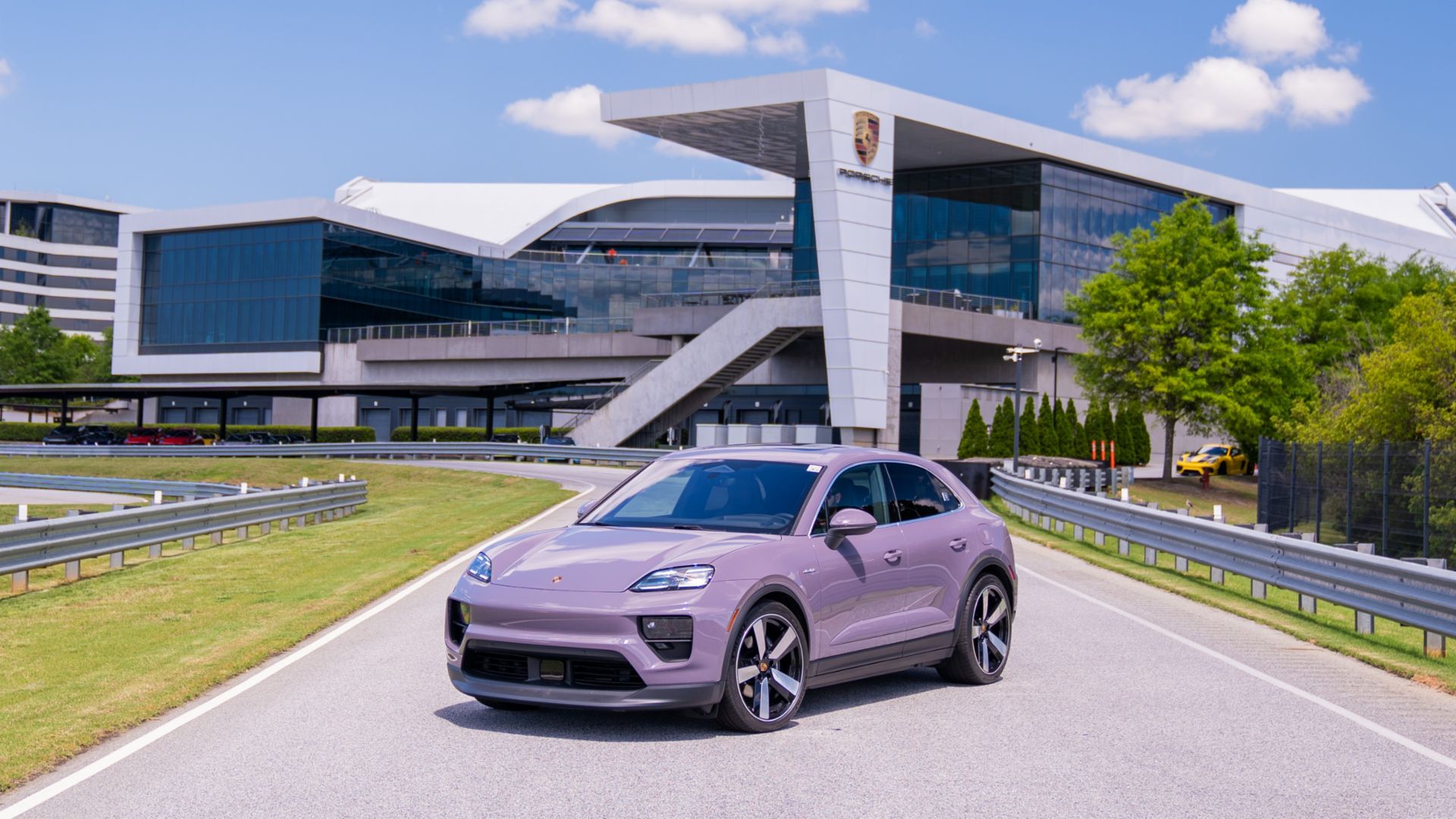 A lavender SUV is parked on a road in front of a modern building with large glass windows and angular architecture. The sky is clear with a few clouds.
