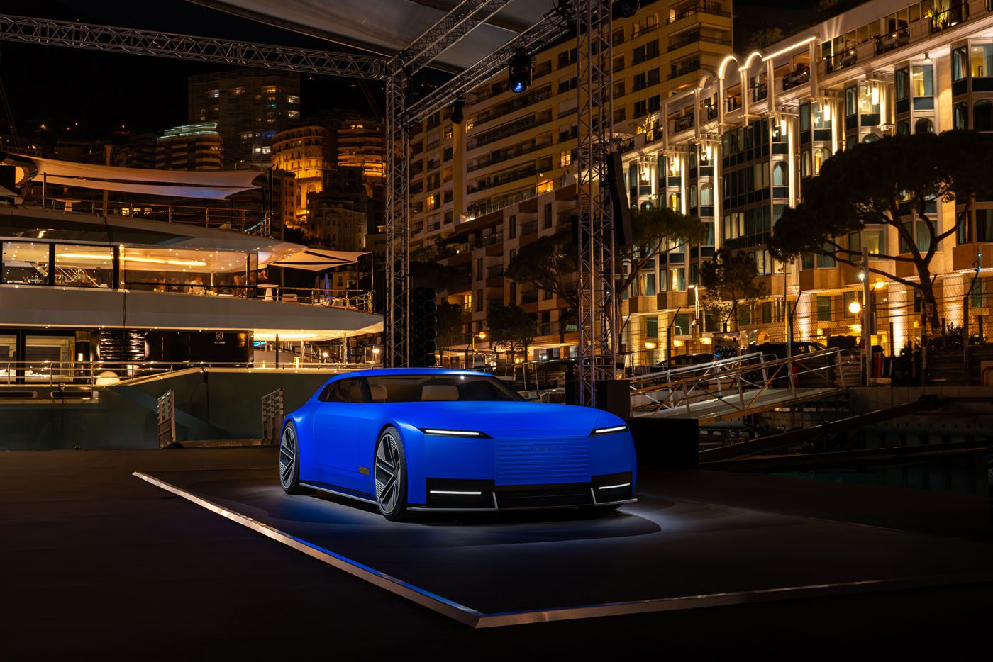 A modern, matte blue concept car is displayed on a lit platform at an outdoor nighttime event with illuminated buildings in the background.