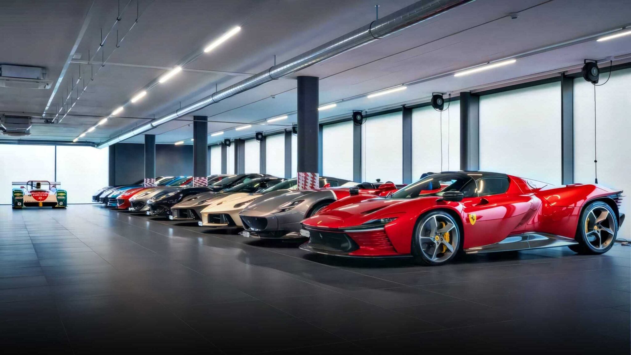 A row of luxury sports cars, including a red hypercar in the foreground, is parked inside a brightly lit, modern showroom with large windows, reminiscent of an RM Sotheby’s Zurich auction display.