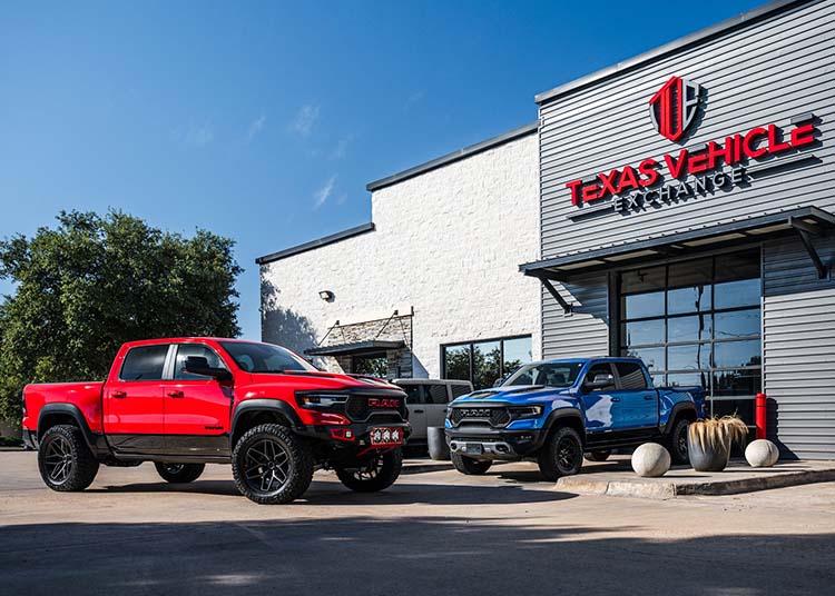 Two pickup trucks, one red and one blue, are parked in front of a building with a sign reading "Texas Vehicle Exchange," known for its selection of custom 4x4s and exotic cars, on a sunny day.