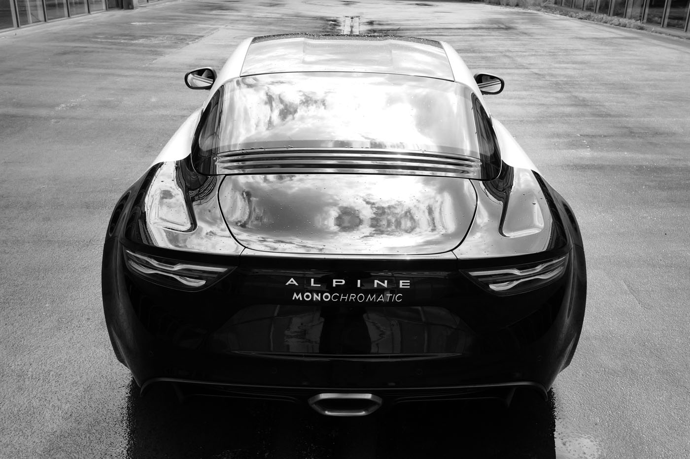 The sleek Black Alpine A110 sports car, an Art Car masterpiece, proudly displays "Monochromatic" on its rear. It's parked on a glistening wet road, viewed from behind in stunning elegance.