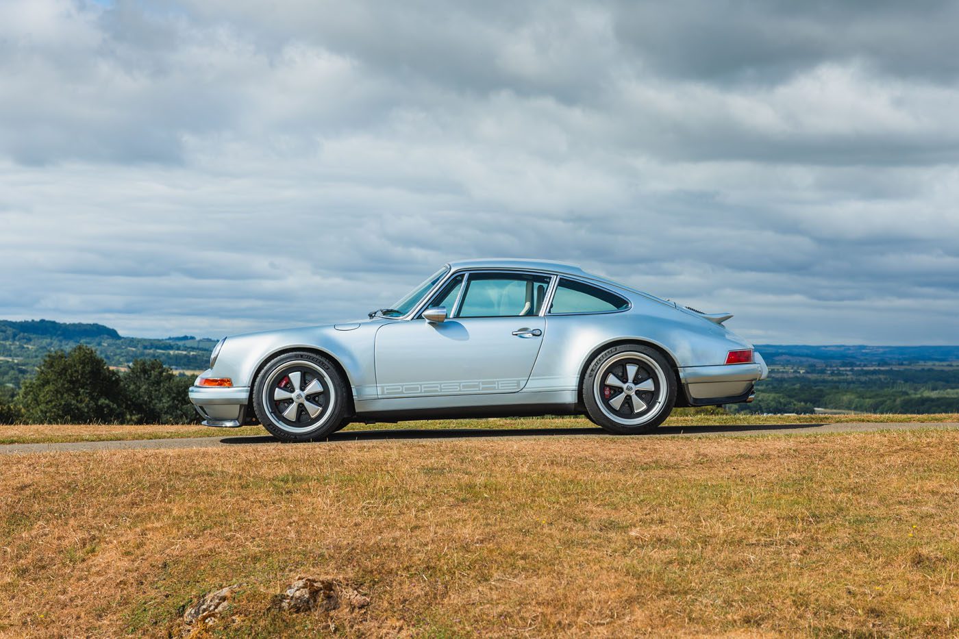 A silver air-cooled 911 coupe by Theon Design is parked on a grassy hill, set against a scenic landscape and cloudy sky.