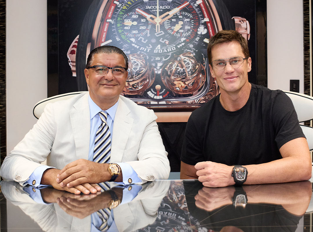 Two men sit at a table in front of a large Jacob & Co. Twin Turbo Furious watch image, both wearing wristwatches and glasses; one wears a suit, the other a black t-shirt.