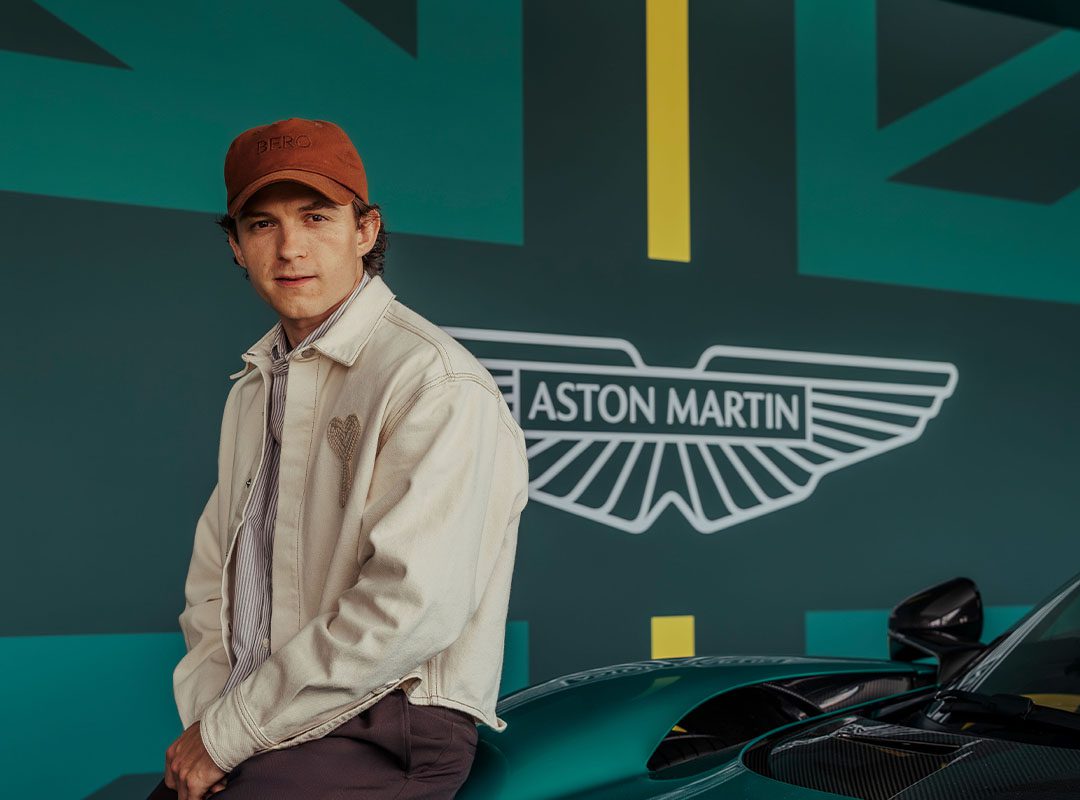 A person wearing a beige jacket and red cap sits on a green Aston Martin car in front of a wall featuring the Aston Martin logo, a stylized British flag design, and the name Tom Holland.