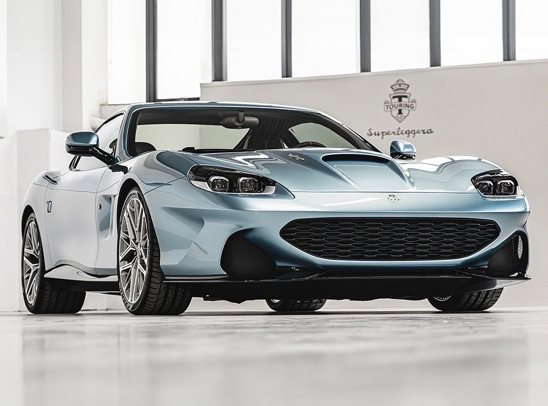 A light blue Touring Superleggera sports car with a sleek design is parked indoors on a white floor at a North American dealership, viewed from a low front angle.