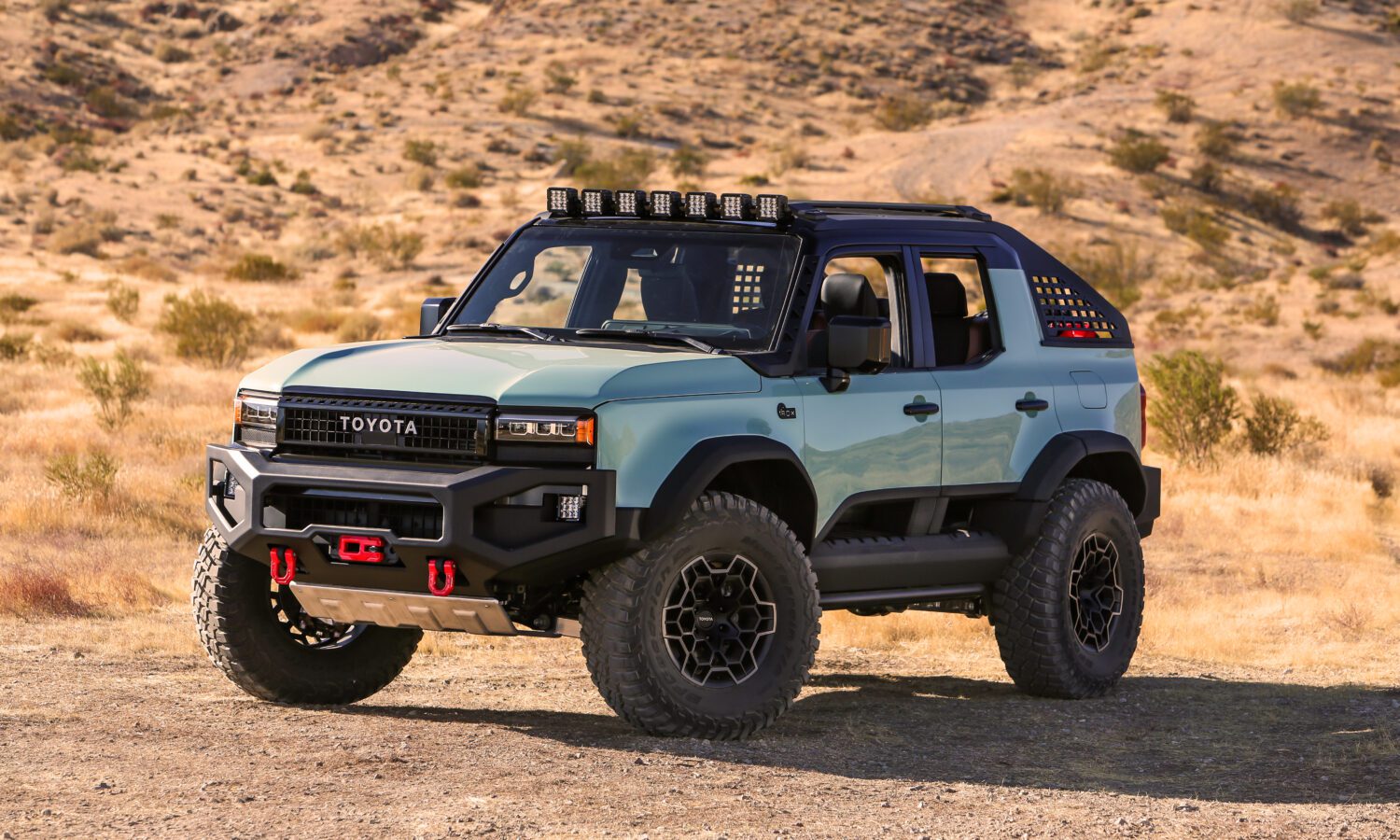 The 2024 SEMA-ready Toyota is a rugged off-road vehicle boasting a striking blue and black exterior. Parked on a dirt path in the dry, hilly landscape, it features roof lights and large tires, perfect for any adventure concept you can imagine.