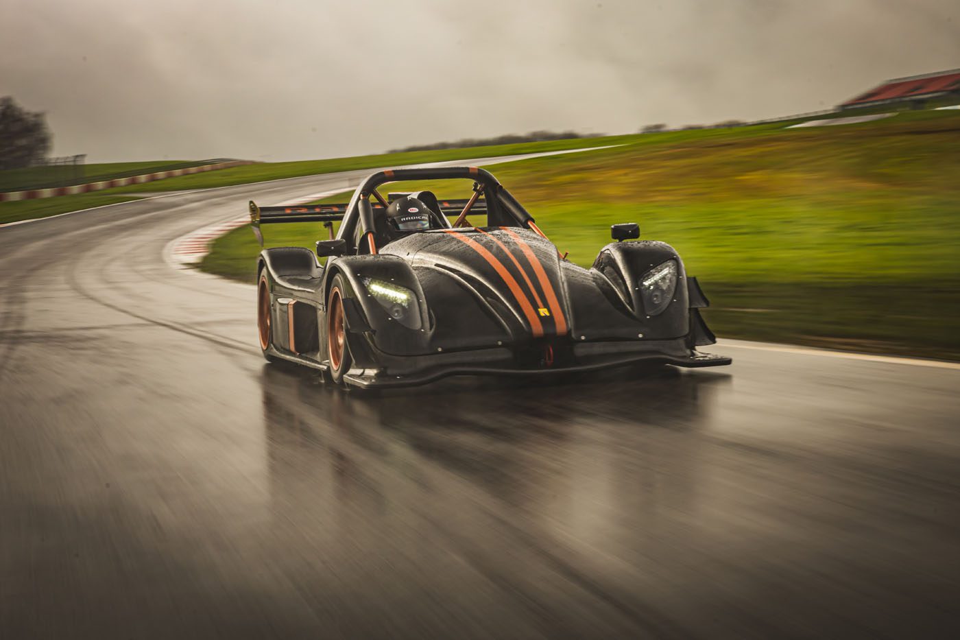 A black race car with orange accents from Radical Motorsports speeds around a wet, curving track under an overcast sky, celebrating the company's 28th Anniversary.
