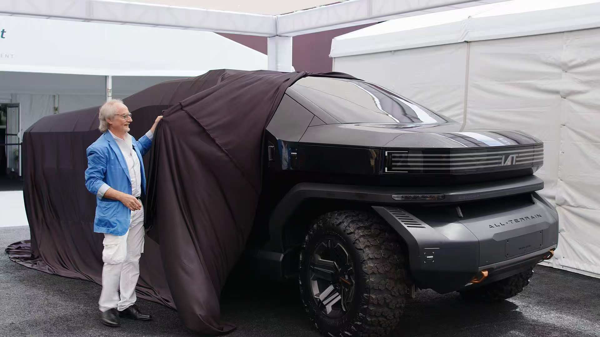 A man in a blue jacket unveils a large black futuristic off-road vehicle partially covered by a dark cloth.