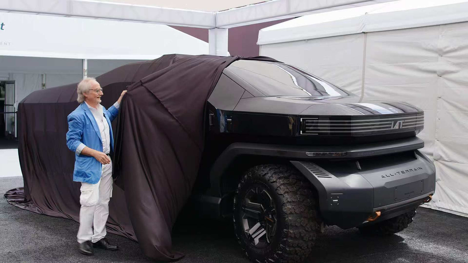 A man in a blue jacket unveils a large black futuristic off-road vehicle partially covered by a dark cloth.