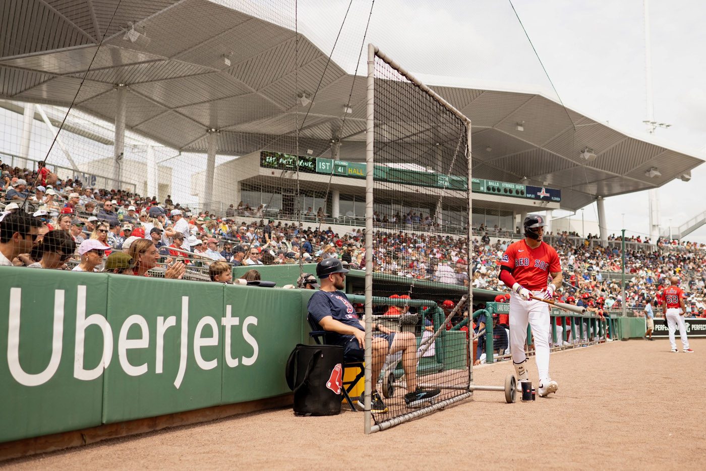 A baseball player stands near the dugout, holding a bat, much like a pilot readying in a virtual hangar. The stadium is filled with spectators watching the game unfold with excitement, reminiscent of an aircraft marketplace buzzing with activity.