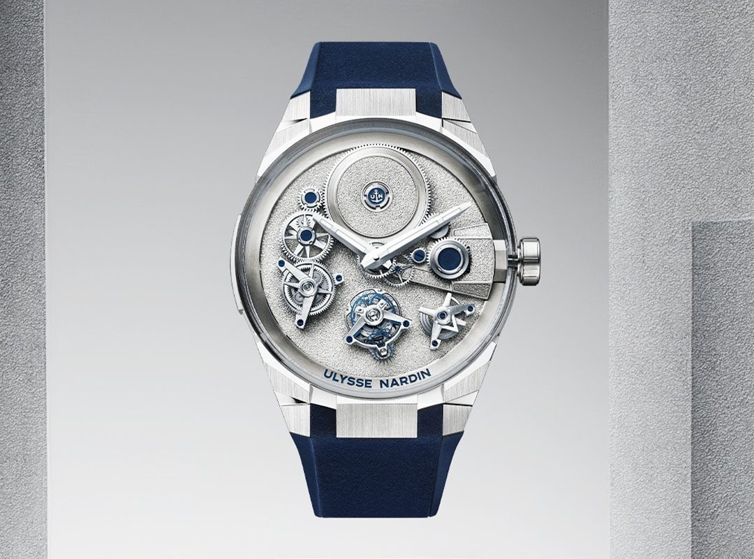 A silver Ulysse Nardin wristwatch with a visible mechanical movement, blue strap, and metallic accents, displayed against a gradient gray background.