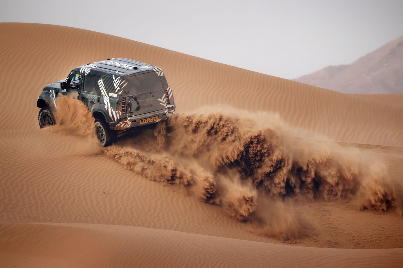A four-wheel drive SUV drives up a sand dune in a desert, kicking up a large spray of sand behind its tires.