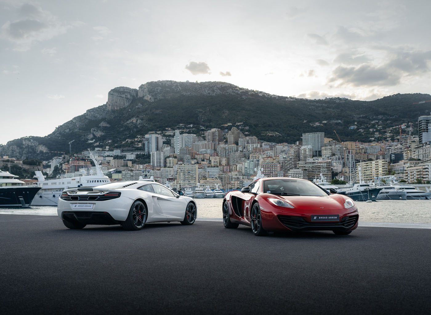 Two sports cars, one white and one red, are parked on a waterfront with yachts and a cityscape backed by hills—evoking the excitement of a McLaren auction and the racing spirit of Lewis Hamilton.