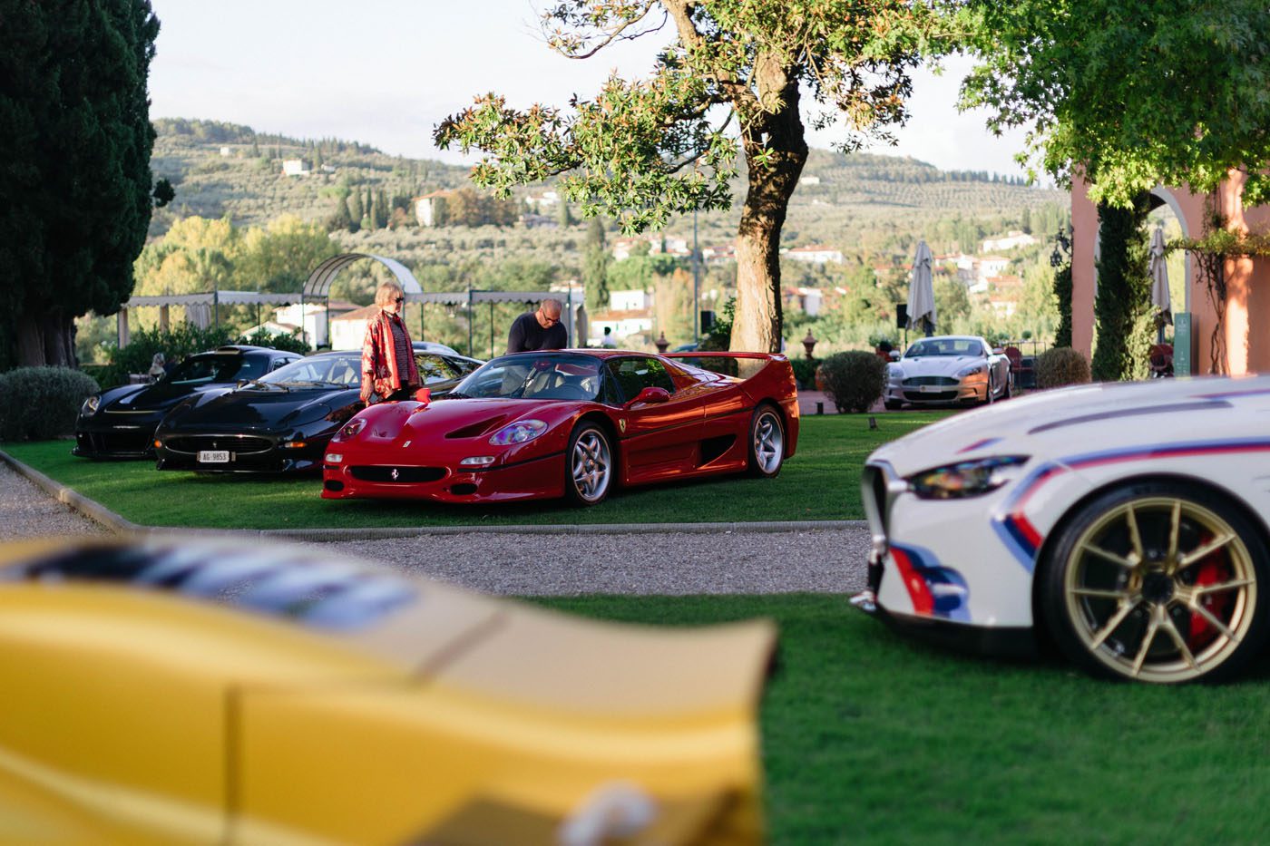 Several luxury sports cars, including a red Ferrari, are parked on a lawn at Villa La Massa, where guests celebrate excellence amid trees and hills. The scene captures the spirit of October 17-19’s exclusive gathering.