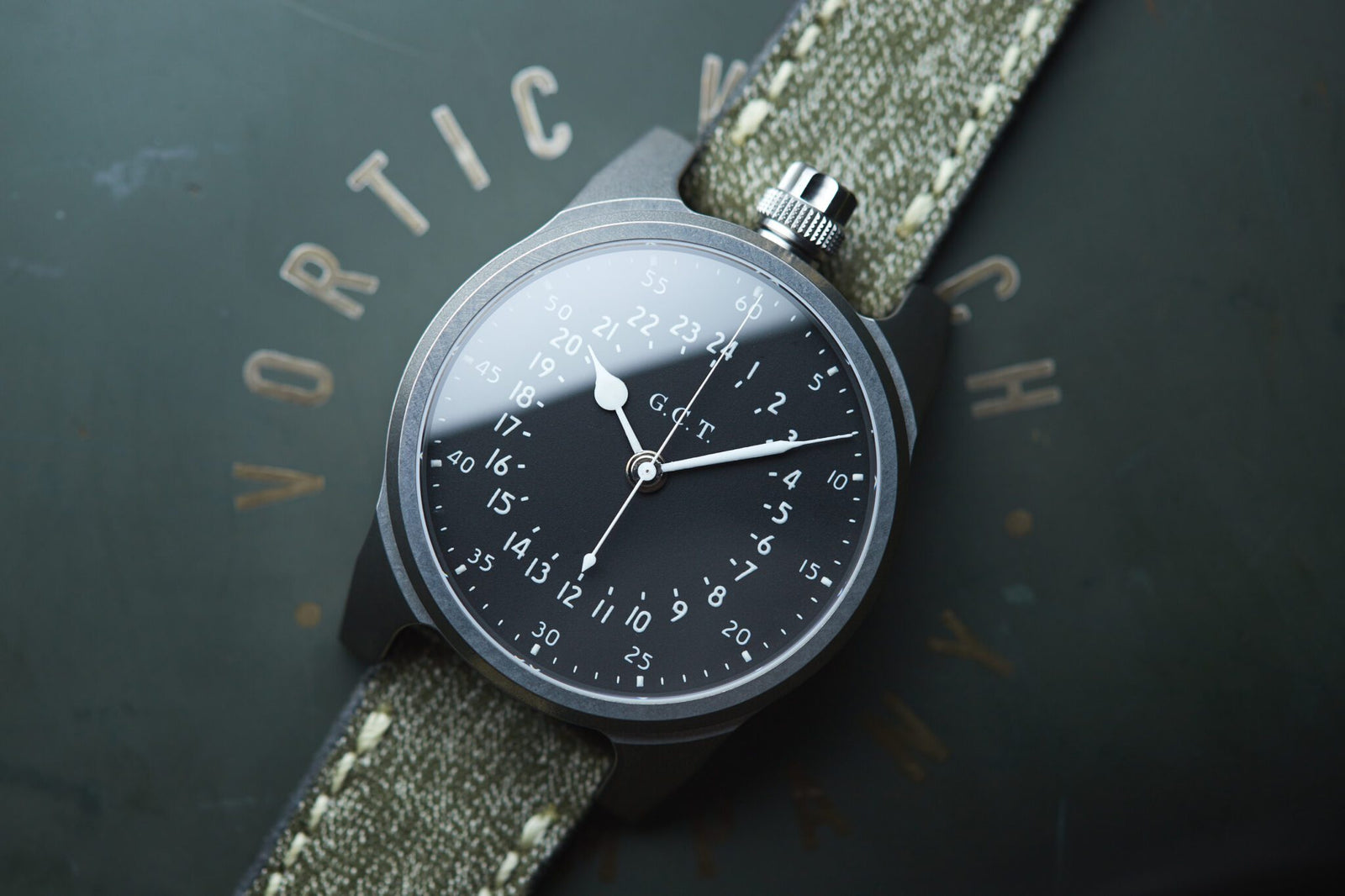 A vintage wristwatch with a dark dial displaying military time numerals and white hands on a green fabric strap lies on a flat surface, showcasing American craftsmanship reminiscent of the Vortic Watch Company's timeless elegance.