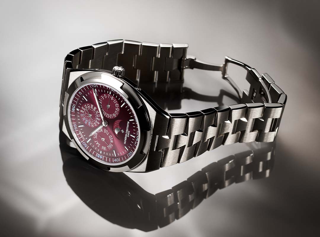 A stainless steel Vacheron Constantin wristwatch with a metallic link band and a burgundy dial featuring multiple subdials, reminiscent of the Overseas Perpetual Calendars, displayed on a reflective surface.