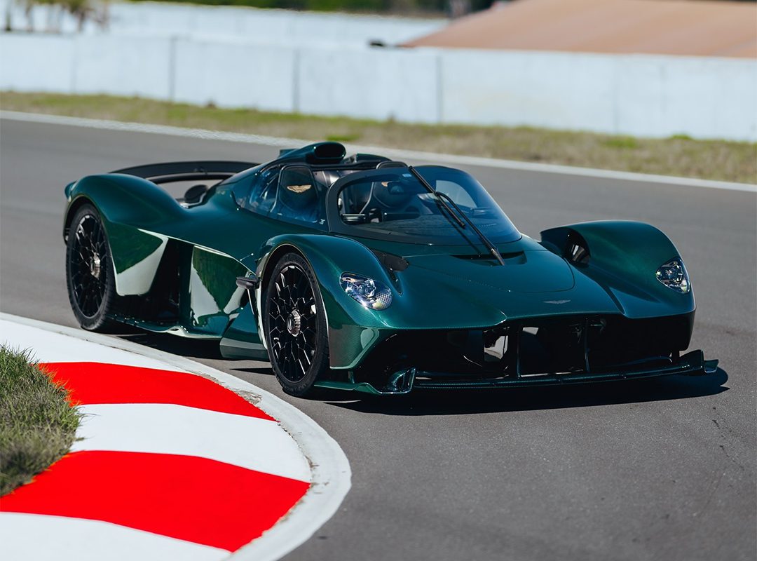 A dark green Aston Martin Valkyrie roadster, with exposed green carbon accents, speeds along a racetrack, rounding a corner as the red and white curb comes into view in the foreground.