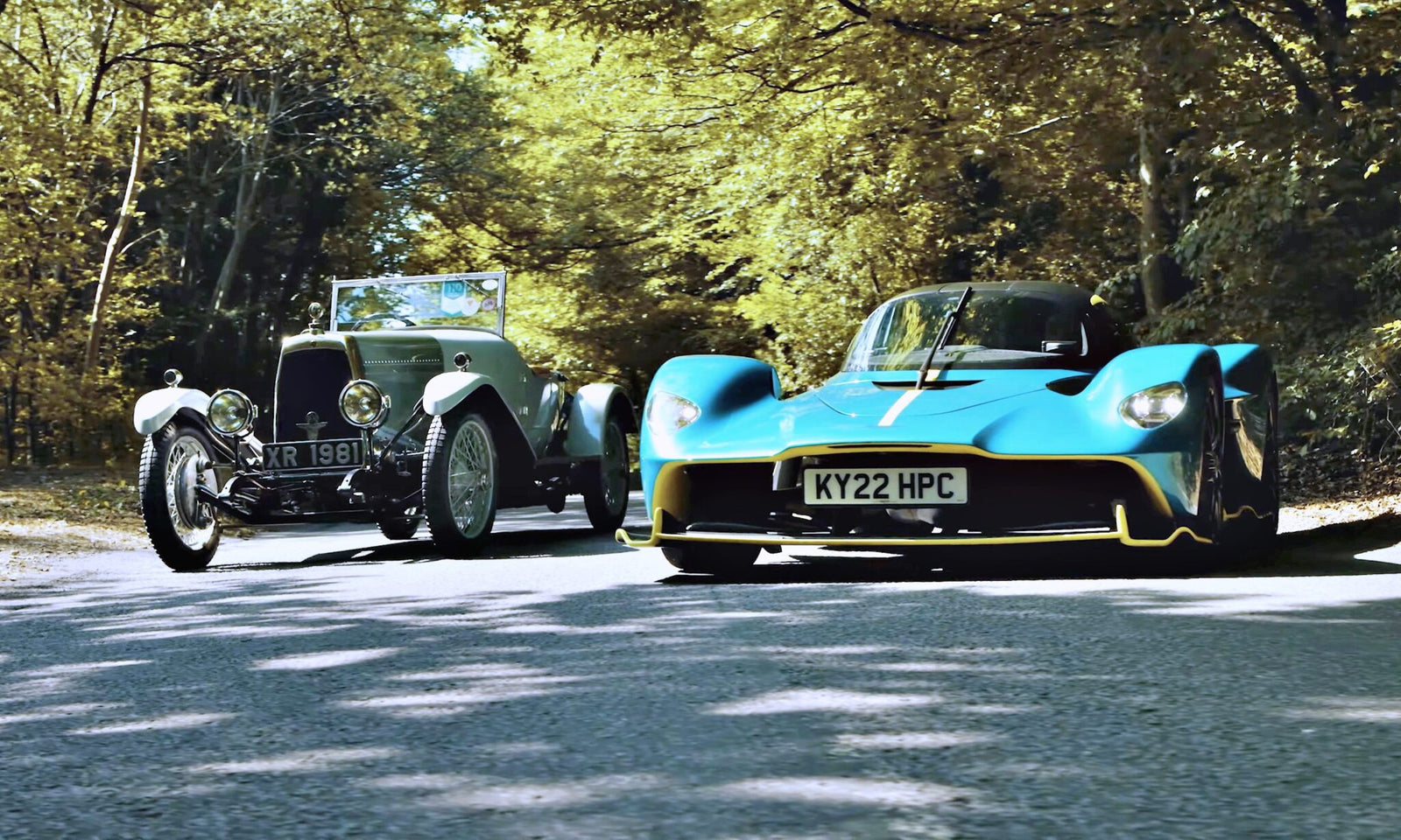 A vintage silver car and a modern blue sports car drive side by side on a tree-lined road in daylight—where Valkyrie Meets Cloverleaf On Aston Hill: Where It All Began 100 Years Ago.