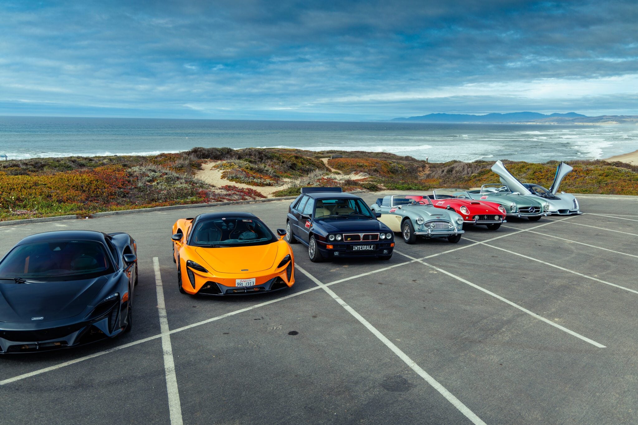 Six cars, including classic and modern models, are parked in a row near a beach with ocean waves and a cloudy sky in the background.