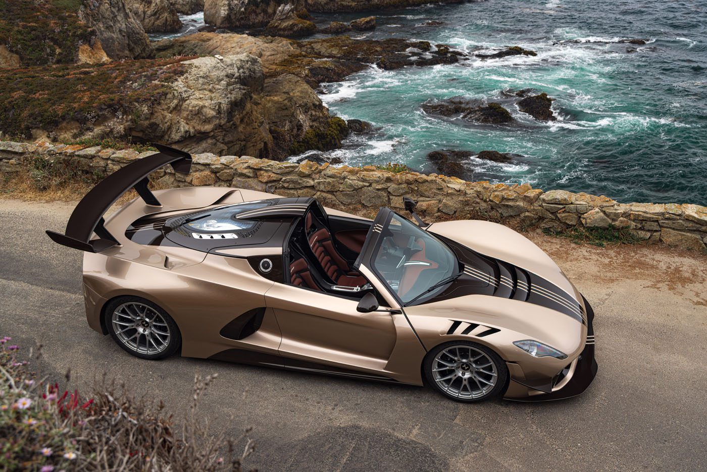 A gold Hennessey Venom F5 convertible with black accents is parked on a coastal road overlooking rocky cliffs and the ocean.