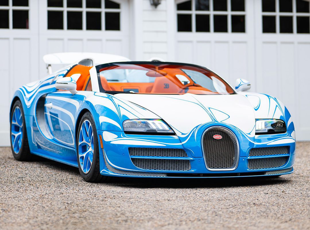 A blue and white Bugatti Veyron Grand Sport Vitesse convertible is parked on a gravel driveway in front of white garage doors.