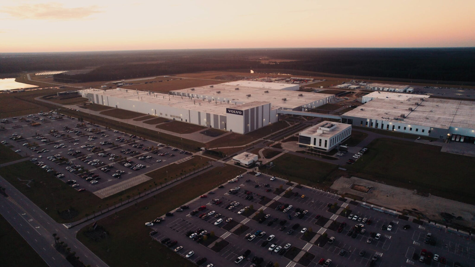 Aerial view of the Volvo South Carolina plant, showcasing multiple buildings, a main entrance, and parking lots filled with cars at sunset—home of the next-gen hybrid XC60.