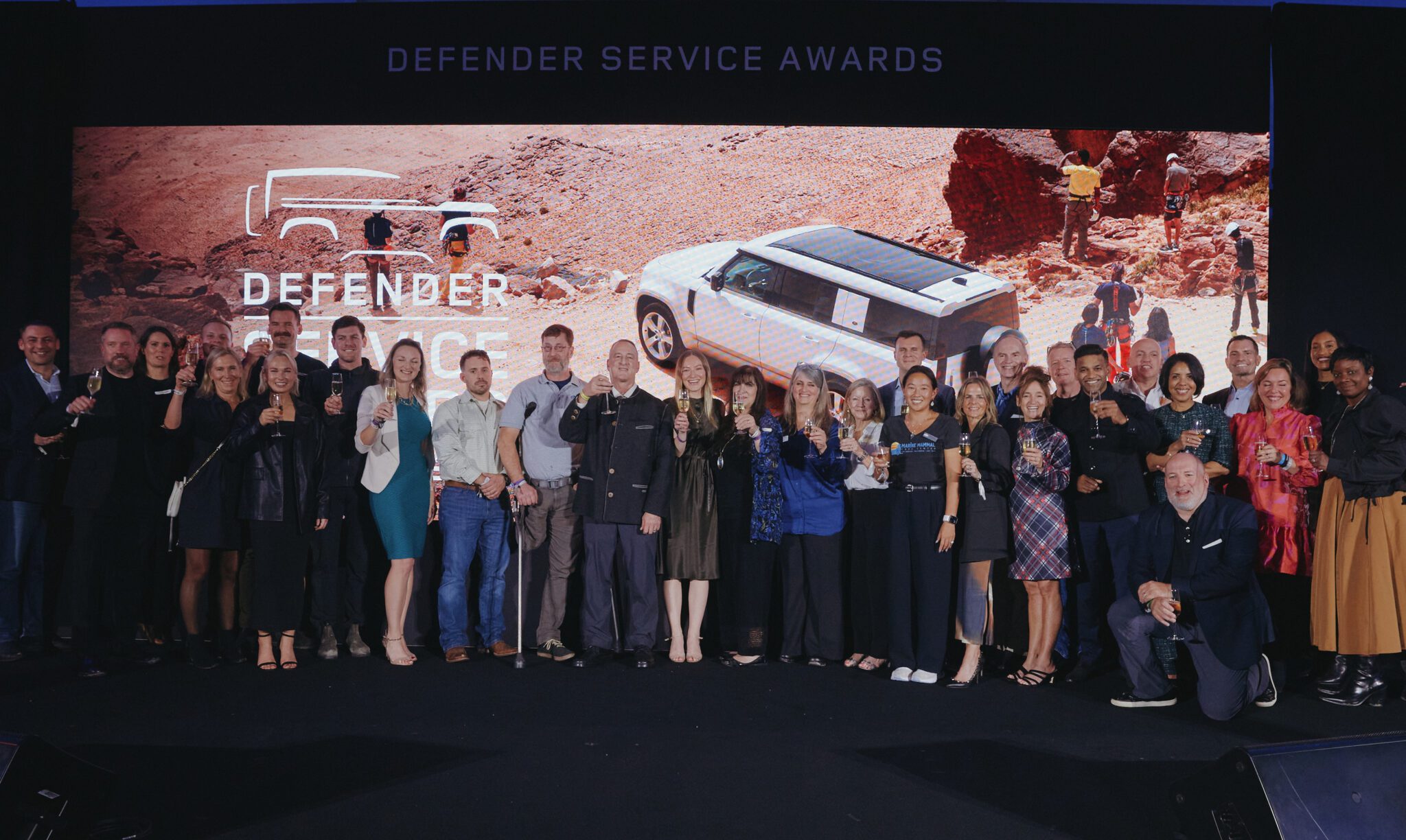 A large group of people pose for a photo on stage in front of a Defender Service Awards sign and an image of a white SUV in a desert landscape.