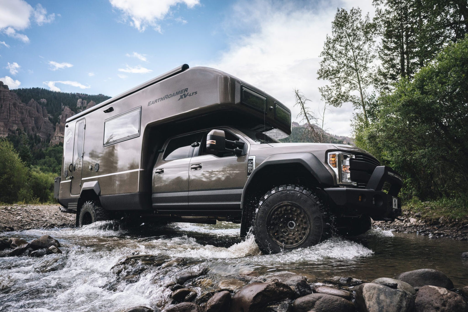 An adventure-ready expedition vehicle drives through a shallow rocky stream, surrounded by trees and mountains under a cloudy sky.