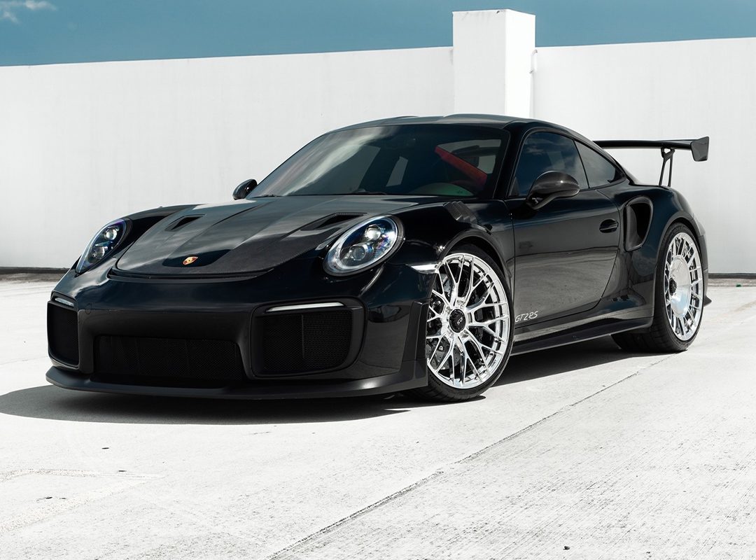 A black Porsche GT2 RS with chrome wheels is parked on a concrete surface against a white wall under a blue sky.