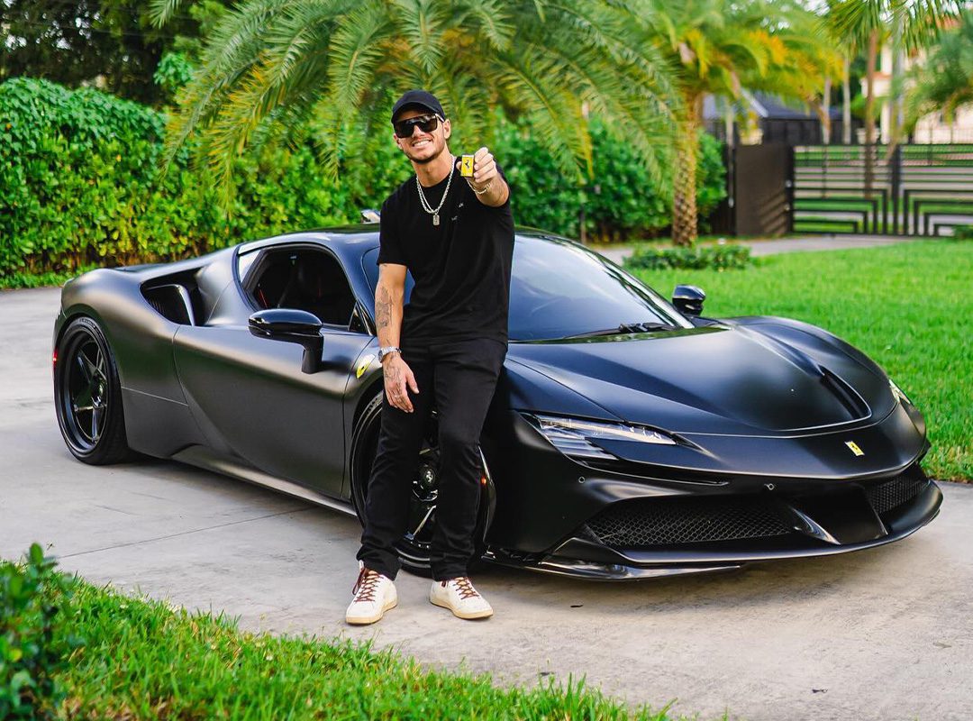 A person in black clothes and a cap sits confidently on a sleek Ferrari SF90 Stradale, giving a thumbs up. The powerful 986HP machine stands out against the lush greenery, capturing the vibe of WhistlinDiesel's adventurous spirit.