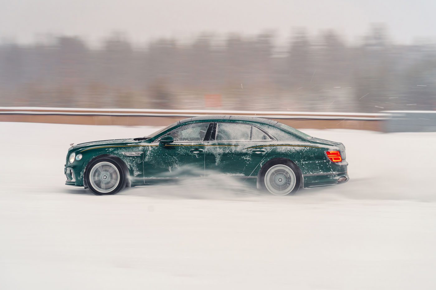 A dark green luxury sedan driving quickly through snow, creating a cloud of snow behind it, with a blurred winter landscape in the background.