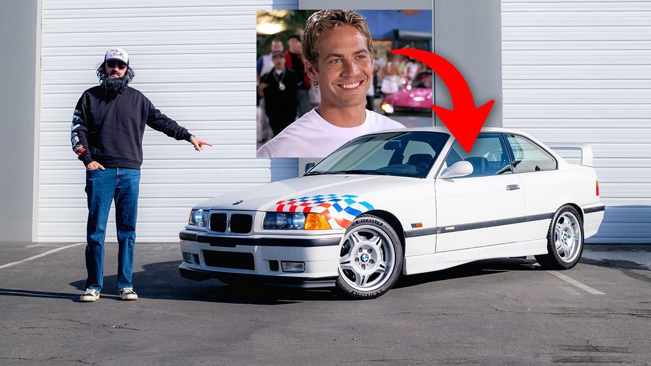 A person stands next to a white E36 BMW M3 Lightweight with a racing flag design. A red arrow points to a photo of another person, possibly Supercar Ron, on the wall behind.
