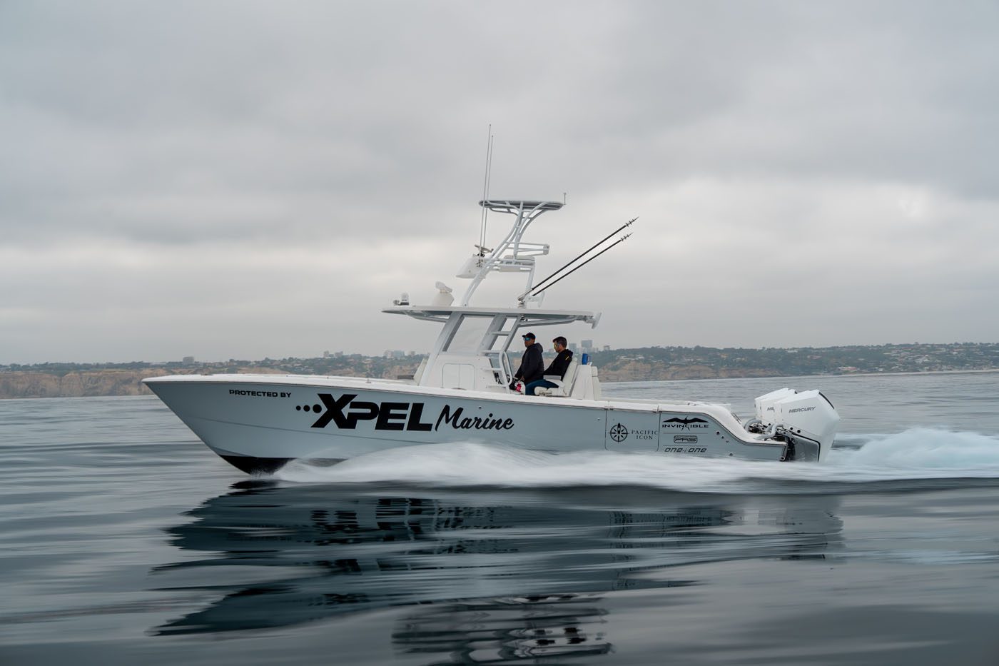 A white motorboat with "XPEL Marine" branding moves across calm water, with two people onboard and a coastline visible in the background under a cloudy sky.