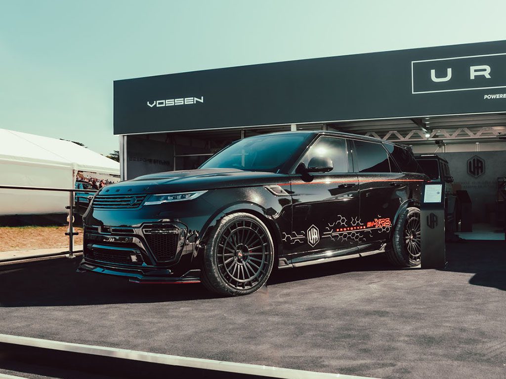 A black Urban Automotive XRS Widetrack SUV with sporty detailing and decals is displayed on a platform at an outdoor auto event at Goodwood, with branded signage in the background.
