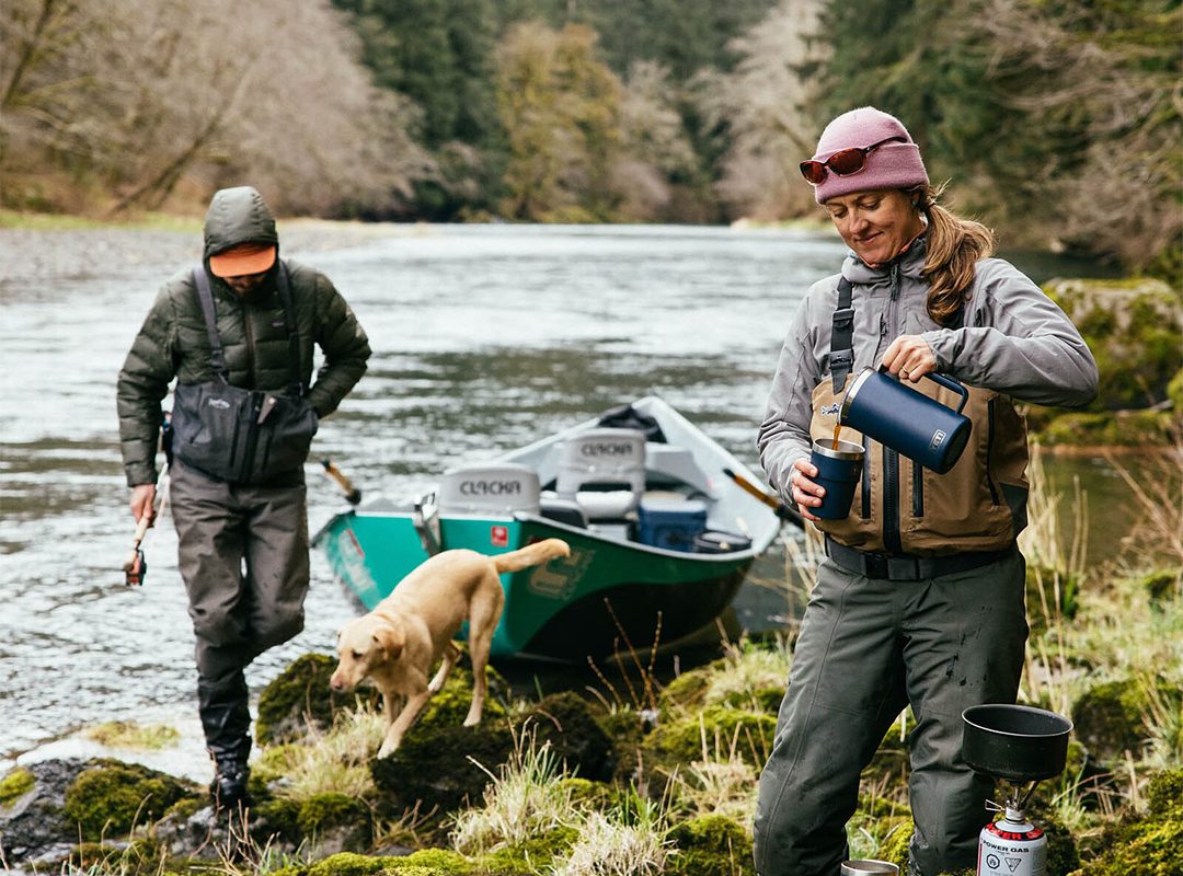 Two people and a dog are near a river with a drift boat; one pours a drink while the other holds a fishing rod. Surrounded by dense forest, they’re ready for an outdoor adventure with their essential gear.