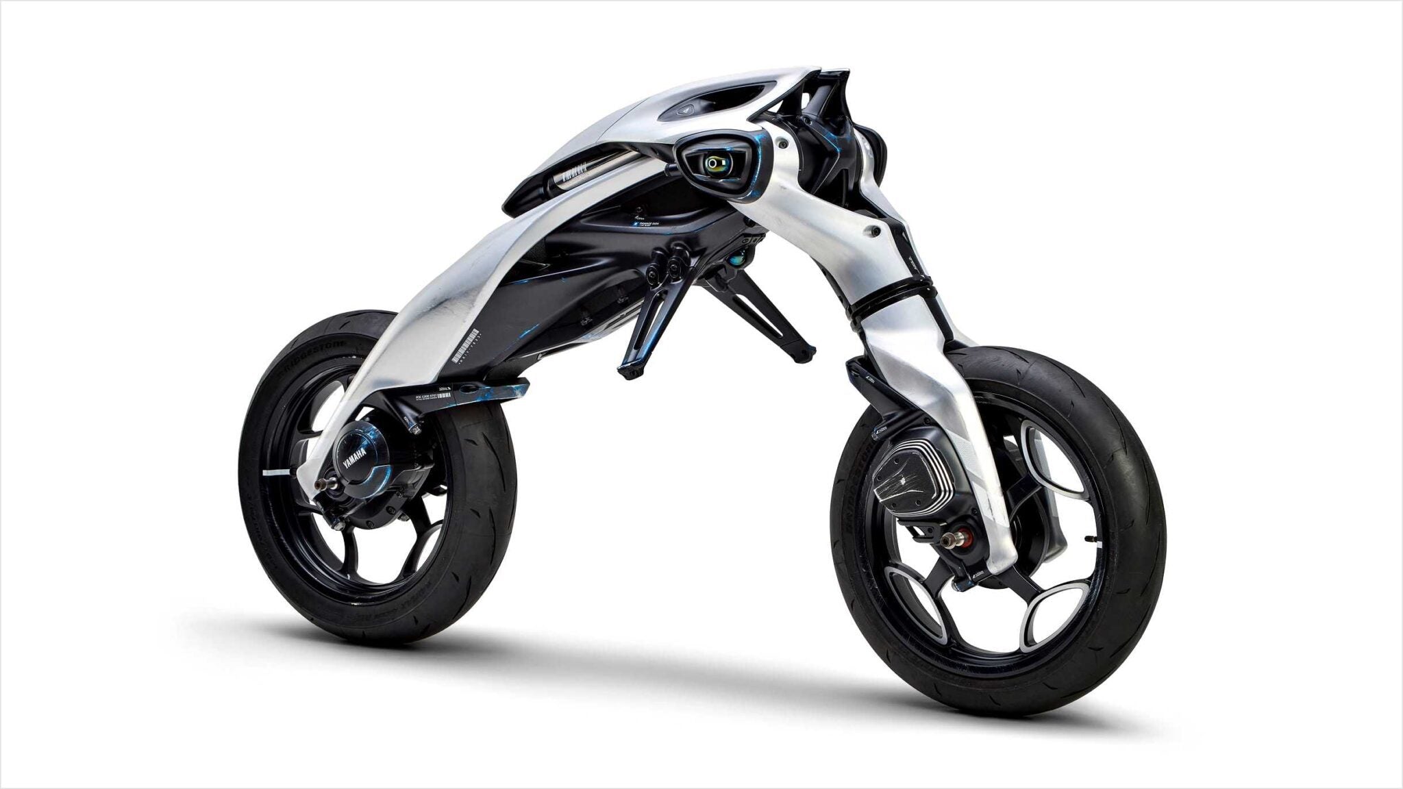 Futuristic, minimalist Yamaha MOTOROiDΛ electric motorcycle with exposed frame, two large wheels, and a sleek, aerodynamic white and black body design against a white background.