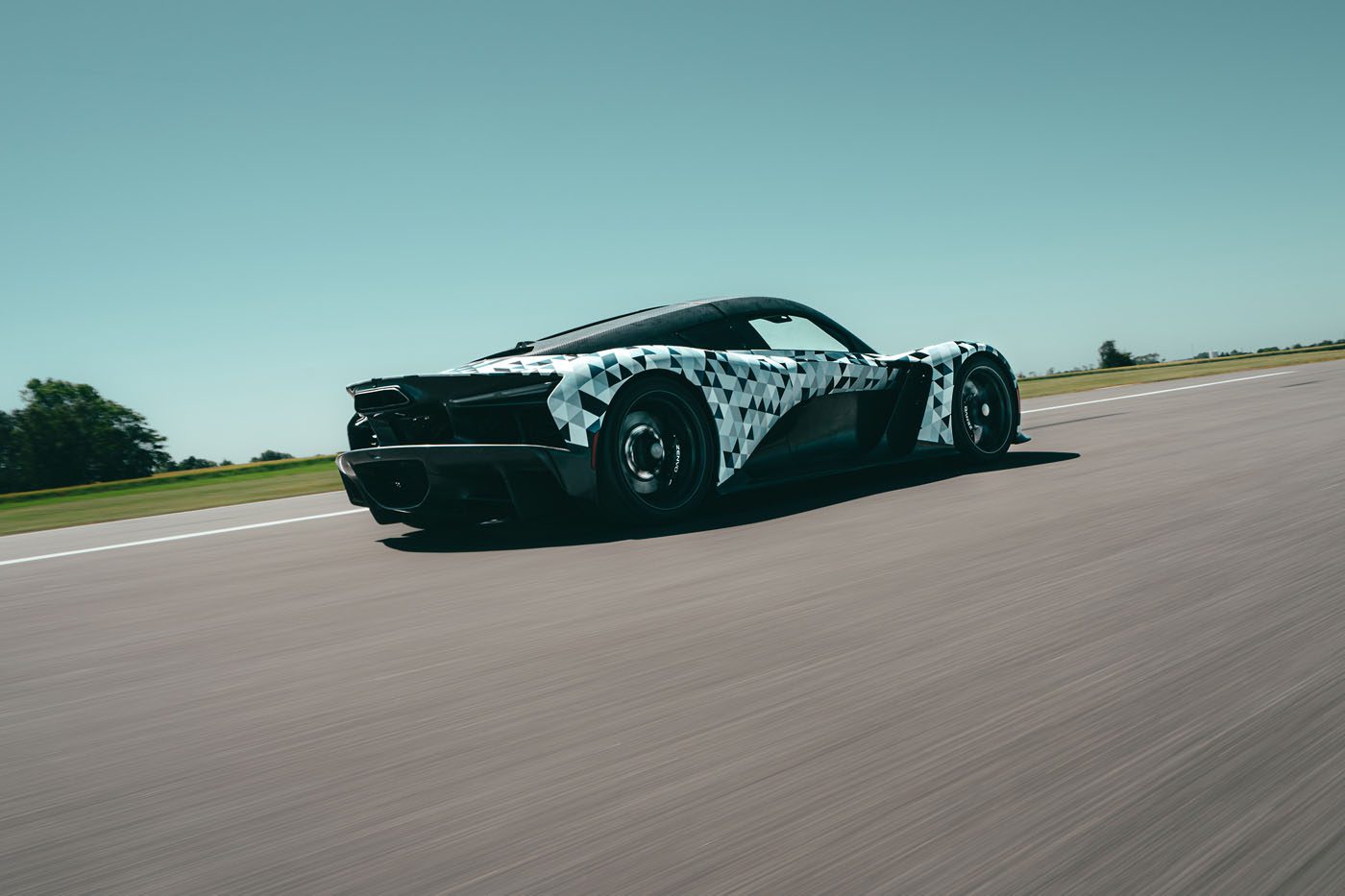 A Zenvo sports car with a black and white camouflage wrap drives at speed on a clear, open road under a blue sky, likely testing ahead of Monterey Car Week as part of the Aurora Development program.